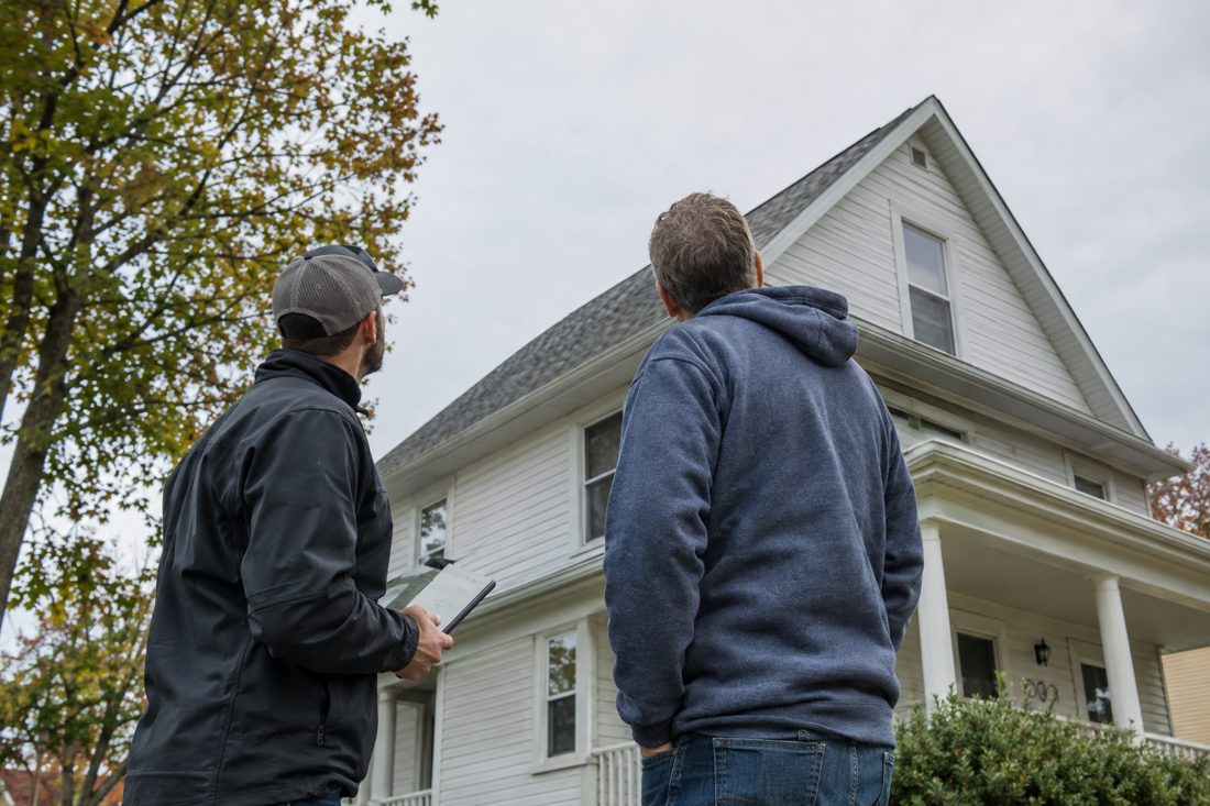 A homeowner and estimator looking up at the roof from the driveway with a tablet at a two-story Iowa farmhouse-style home with maples in Waterloo, IA