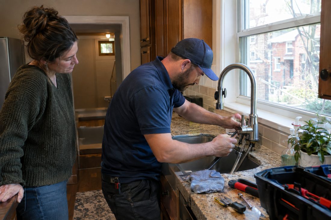 A plumber replacing a kitchen faucet with a homeowner watching at a red-brick Cincinnati home with a basement in Cincinnati, OH